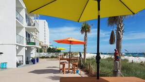 A beachside patio with wooden tables and benches, yellow and orange umbrellas, and a view of the beach and ocean.