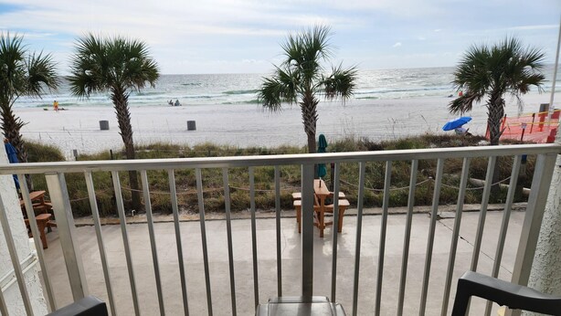 A balcony with two chairs, a table, and a view of the beach with palm trees and the ocean. - Ambassador 126 - Gulf Front (Panama City Beach)