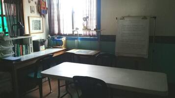 Un aula con una mesa larga, sillas, una pizarra y un ventanal con cortinas.