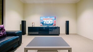 Ett modernt vardagsrum med en platt-TV på ett TV-bord, ett svart soffbord och en svart lädersoffa med en lila prydnadskudde.