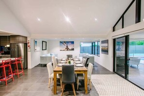 A modern dining area with a wooden table and chairs, a kitchen with a refrigerator and stove, and large windows offering a view.