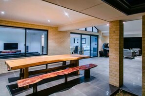 A modern interior with a large wooden table and benches, a brick wall, and a view through a glass door. - Fairways: Spacious Pool House by the Golf Course (Dunsborough)