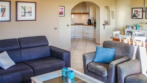 Un salon avec un canapé gris foncé, un fauteuil gris et une table basse en verre agrémentée d’objets décoratifs. On aperçoit une cuisine à l’arrière-plan, avec des meubles blancs et une cuisinière à gaz.