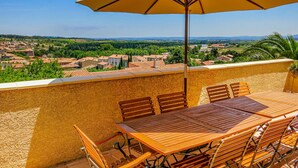 Une terrasse avec une table et des chaises, un grand parasol et une vue sur une ville avec ses maisons et sa verdure.