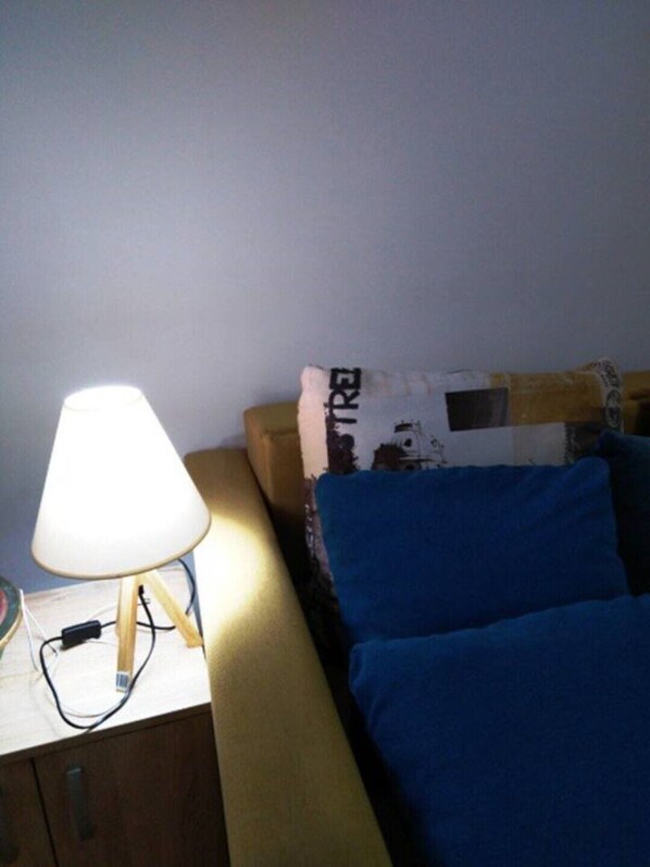 Un lit avec une literie bleue, une lampe sur une table de chevet et un oreiller sur lequel on peut apercevoir partiellement un mot.