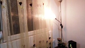 Una habitación con ventana cubierta por una cortina translúcida, una lámpara de pie, un ordenador sobre un escritorio y una cama con cubrecama azul.