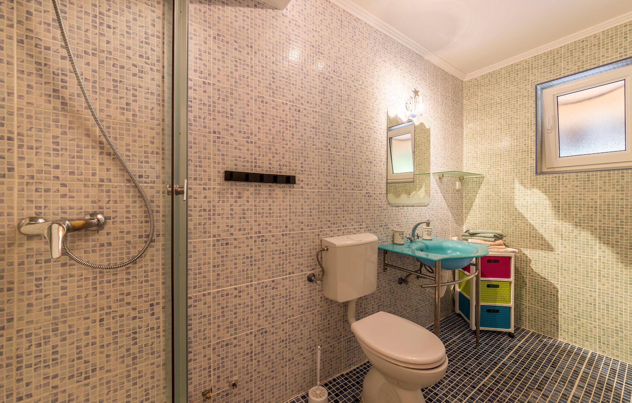 Een badkamer met mozaïek betegelde wanden, een glazen douchecabine, een toilet en een wastafel met een blauwe wasbak.