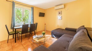 Een woonkamer met een bank, een eettafel met stoelen, een aan de muur bevestigde televisie en een airconditioning.