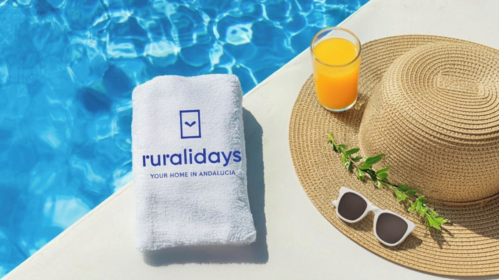 A towel with 'ruralidays' branding, a straw hat, sunglasses, a glass of orange juice, and a pool.