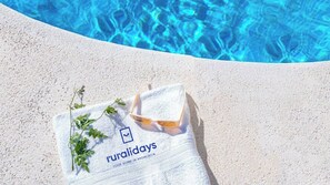 Une serviette ornée du mot « ruralidays » et une plante sur une surface sablonneuse, près d’une piscine.