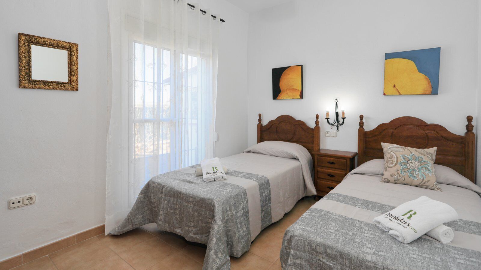 Habitación de hotel con dos camas, cabecera de madera, mesita de noche, lámpara y ventana con cortinas traslúcidas.