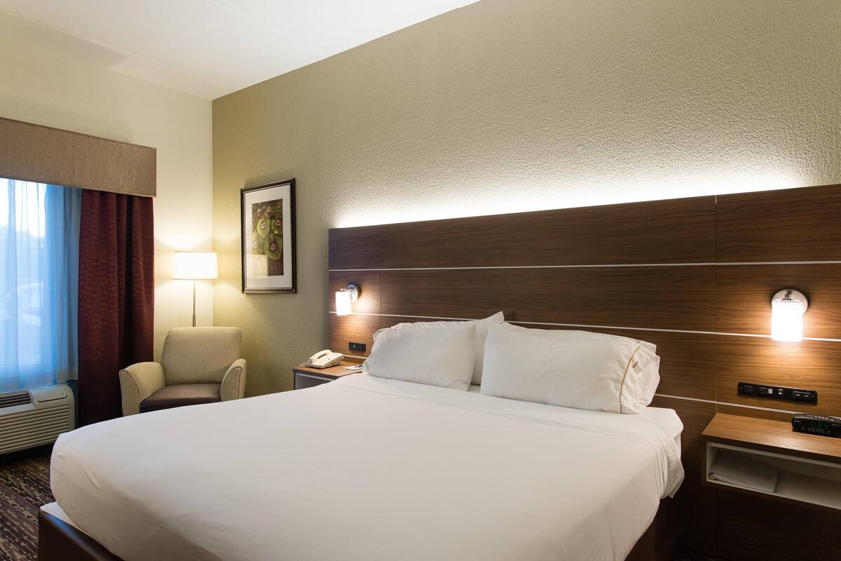 A hotel room with a large bed, a chair, a desk with a lamp, and a nightstand with a phone and a remote control.