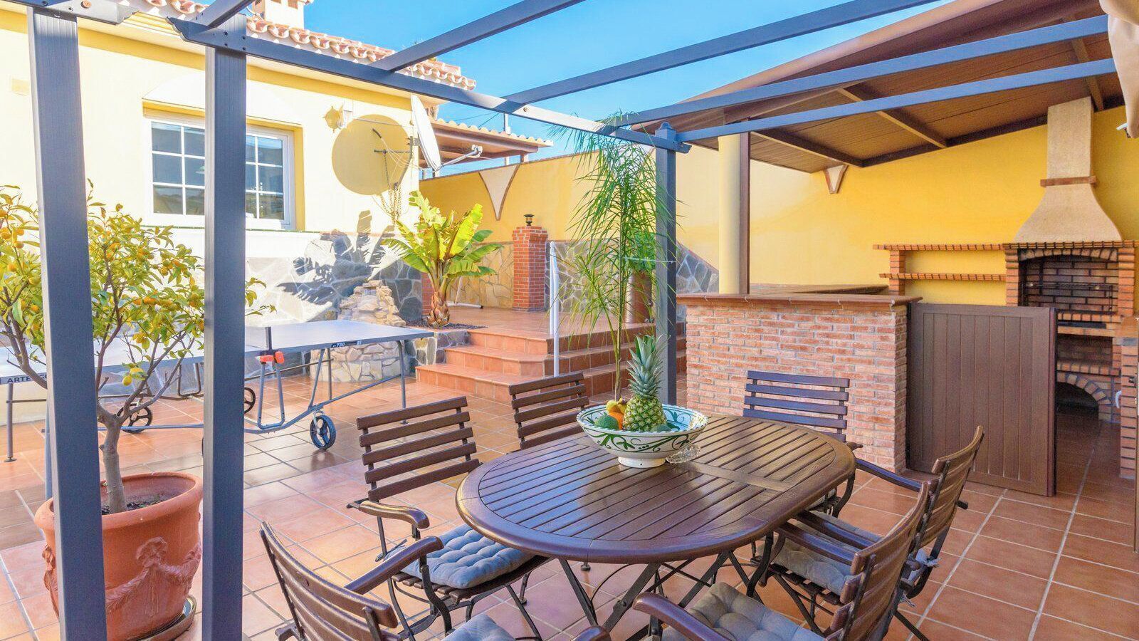 An outdoor patio with a round wooden table and chairs, a potted plant, and a brick oven.