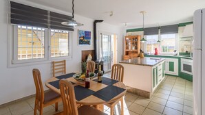 Zona de menjador amb una taula per a quatre, cuina amb armaris verdosos i vista cap a l'exterior a través d'una finestra amb persianes.