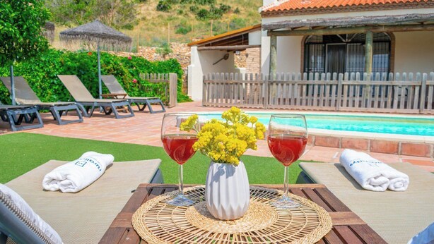 Ein Poolbereich mit zwei Liegestühlen, einem Korbstuhl-Tisch mit einer Vase voller gelber Blumen und zwei Gläsern roter Getränke.