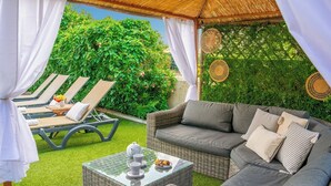 Ein überdachter Außenbereich mit einem Korbsessel, Loungestühlen und einem Tisch mit Teekanne und Tassen.