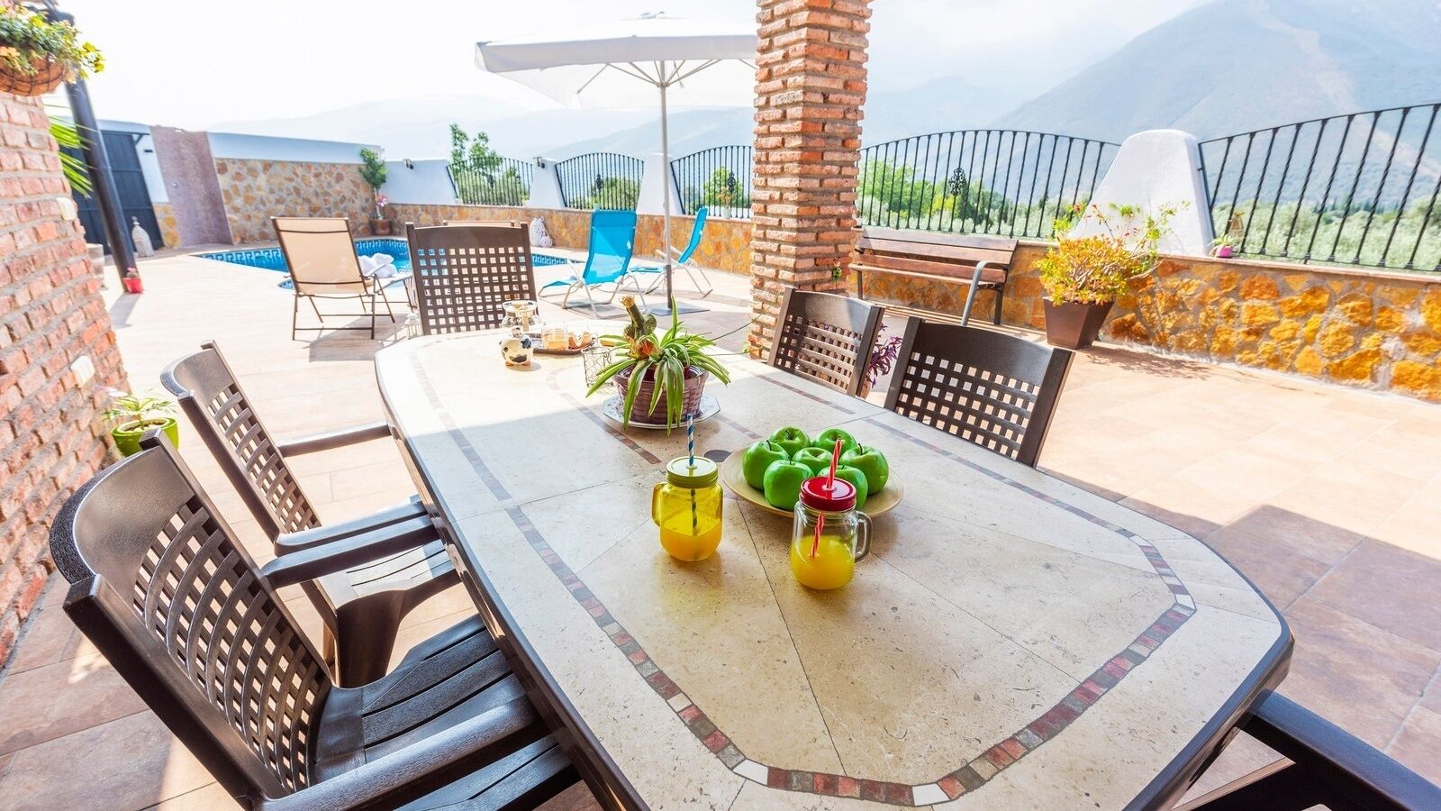 A patio with a table set for dining, surrounded by chairs, with a pool and mountains in the background.