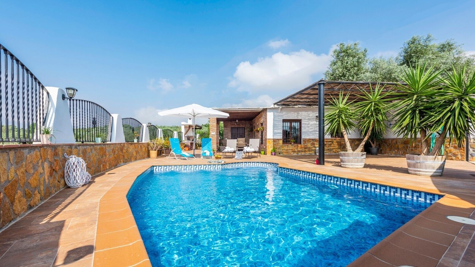 En swimmingpool med liggestole, en stenvæg og en overdækket terrasse.