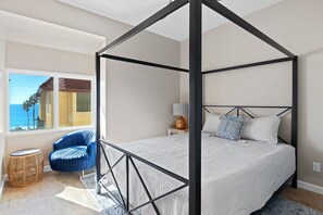 Une chambre à coucher avec un lit à baldaquin, une vue sur l’océan par une grande fenêtre, un fauteuil bleu et une petite table ronde.