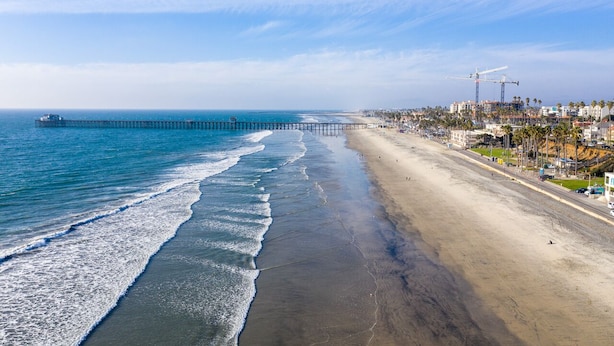 Una playa costera con un muelle que se adentra en el océano, una orilla arenosa y edificios a lo largo de la costa.
