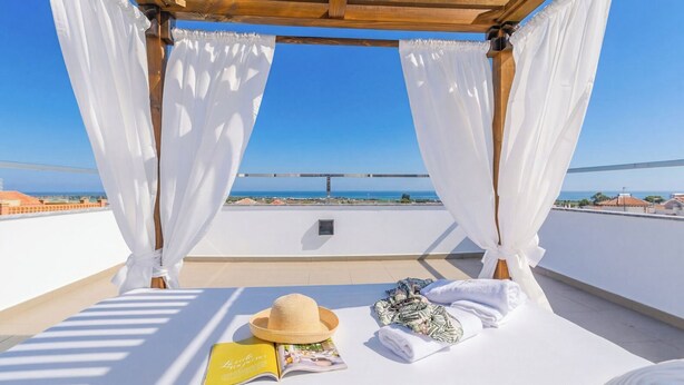 Un balcón con una cama, cortinas blancas, un sombrero de paja y una revista.