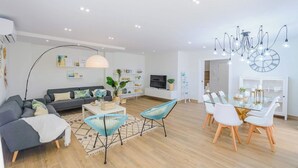 Una sala de estar moderna con sofá, mesa de centro y sillones, un comedor con mesa y sillas de vidrio, y un televisor montado en la pared.