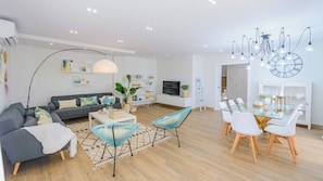 Una sala de estar moderna con sofá, mesa de centro y sillones, un comedor con mesa y sillas de vidrio, y un televisor montado en la pared.