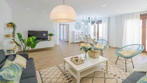 Una sala de estar moderna con sofá, mesa de centro y sillas. Se observa un televisor, una planta y una araña.