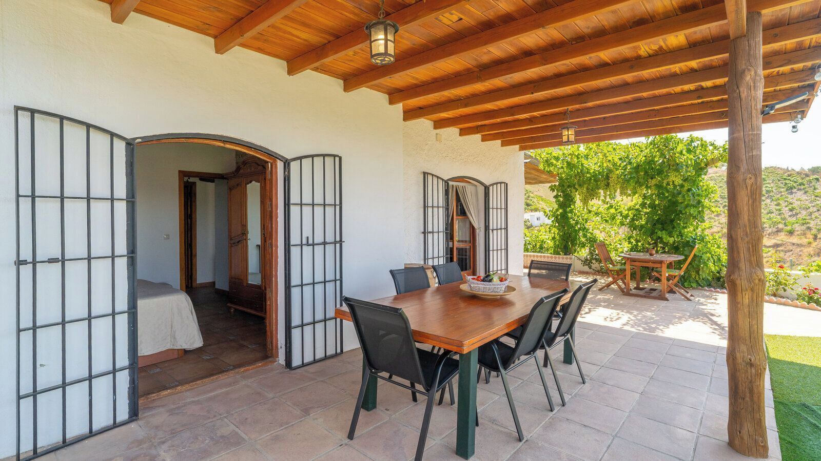 A covered patio with a wooden table and chairs, a view of greenery, and a door leading to a room.