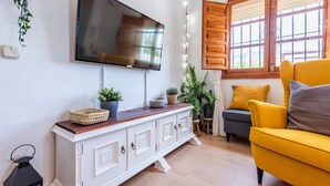 A living room with a flat-screen TV mounted on the wall, a white cabinet with decorative items, a yellow armchair, and a window with wooden shutters. - El Llano del Torcal Antequera - La Higuera by Ruralidays (Málaga)