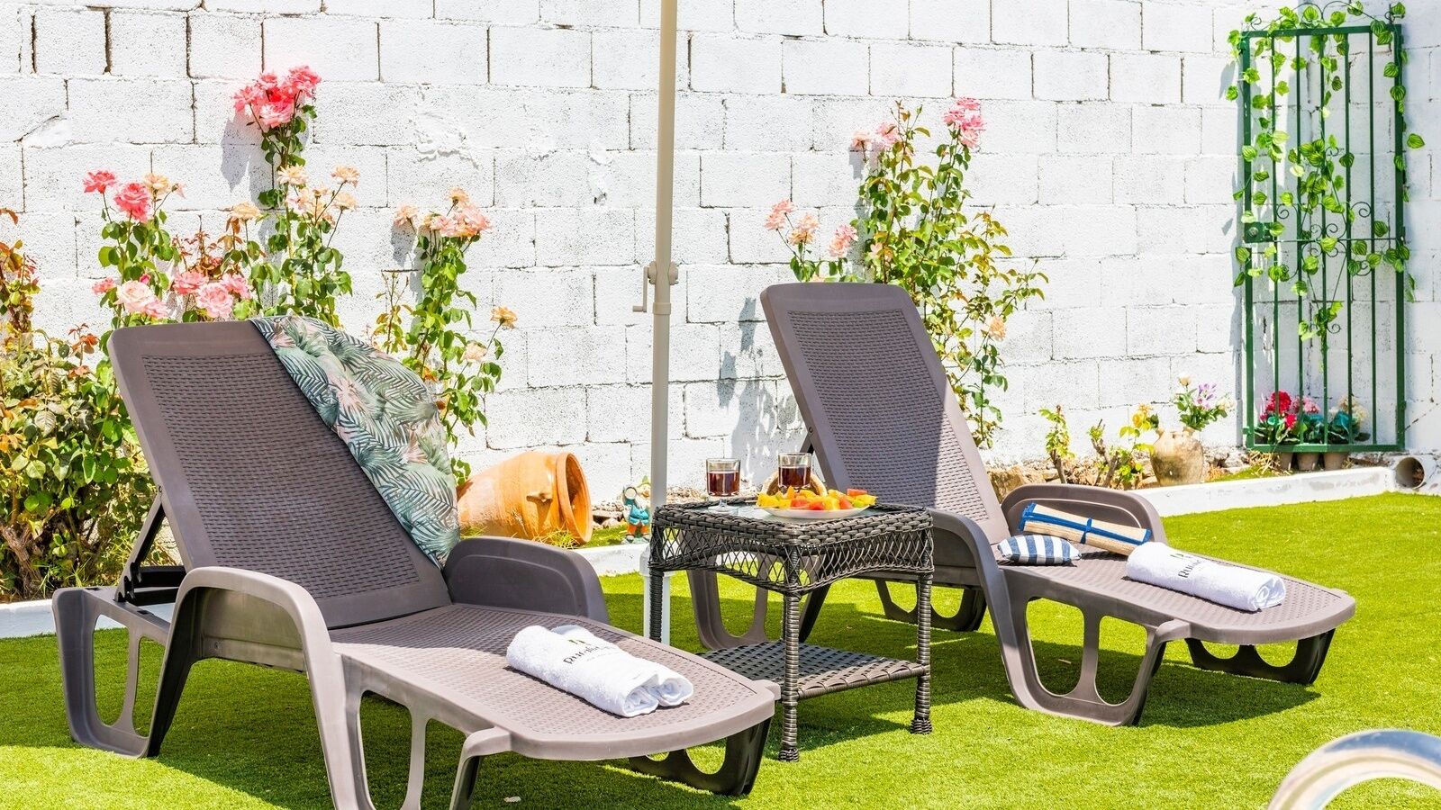 Zwei Liegestühle mit Handtüchern, ein kleiner Tisch mit Getränken und eine weiße Backsteinwand mit Blumen im Hintergrund.