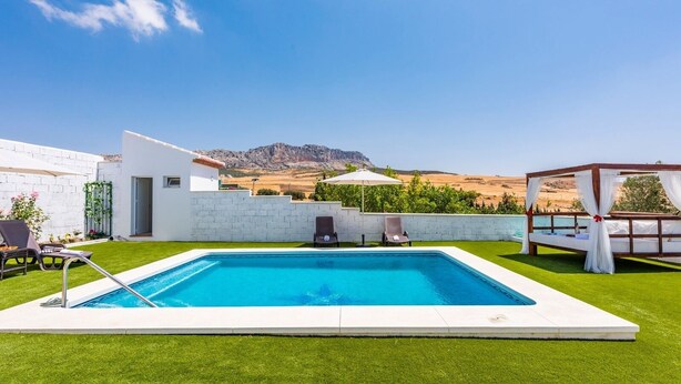 A rectangular swimming pool with a white deck, surrounded by a well-maintained lawn. There are lounge chairs and a pergola with white curtains. - El Llano del Torcal Antequera - La Higuera by Ruralidays (Málaga)