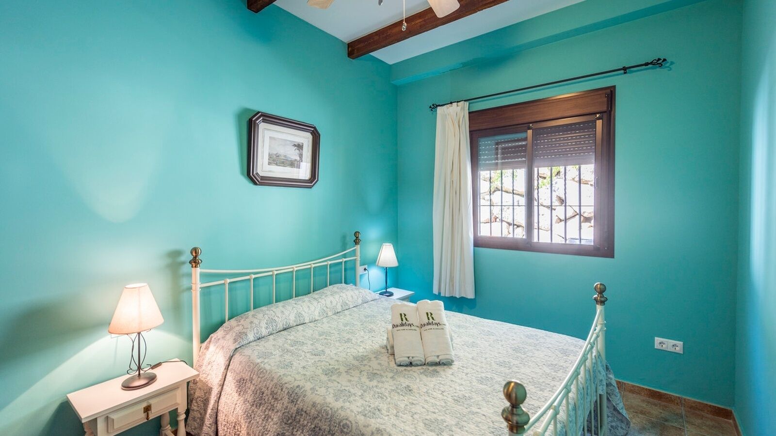 Quarto com cama, mesa de cabeceira, luminária, janela com cortinas e quadro na parede.
