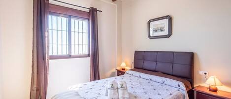 A bedroom with a bed, two bedside lamps, a window with curtains, a framed picture on the wall, and a bedside table.