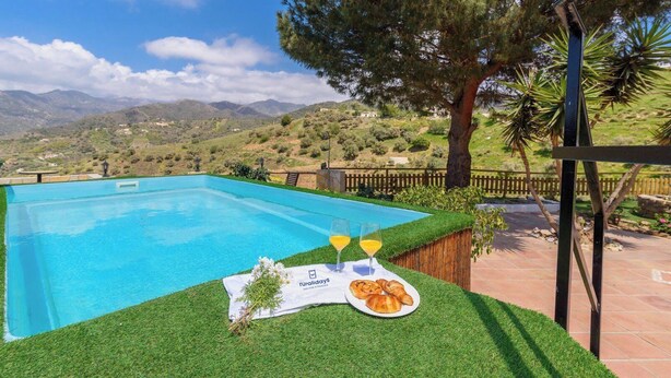 A swimming pool with a grassy area, a table with food and drinks, and a view of mountains in the background. - Cortijo Baeza Torrox by Ruralidays (Málaga)