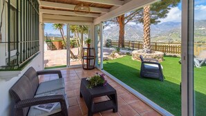 A covered patio with wicker furniture, a small table with a plant, and a view of a landscaped garden with a stone wall and trees. - Cortijo Baeza Torrox by Ruralidays (Málaga)