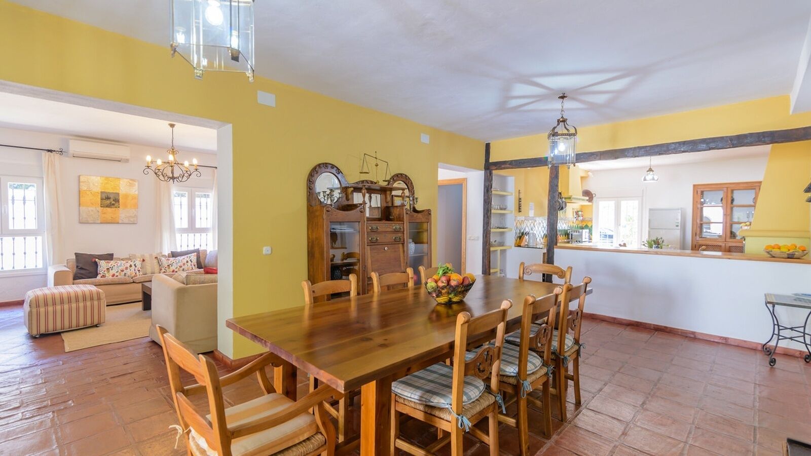 A spacious dining area with a wooden table and chairs, a kitchen in the background, and a living room with a sofa and ottoman.