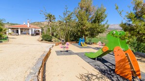 A playground with a slide and swings, surrounded by sand and greenery, with a building in the background. - Casa Aranda Villanueva de la Concepción by Ruralidays (Málaga)
