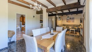 A dining area with a wooden table and white wicker chairs, a chandelier, and a kitchen in the background. - Casa Aranda Villanueva de la Concepción by Ruralidays (Málaga)