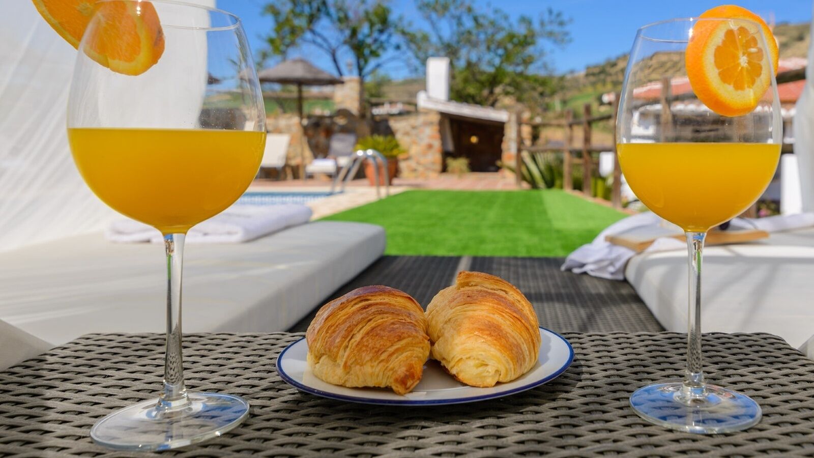 Zwei Croissants auf einem Teller, zwei Gläser Orangensaft und eine Poolanlage mit freiem Blick auf den Pool und ein Gebäude im Hintergrund.