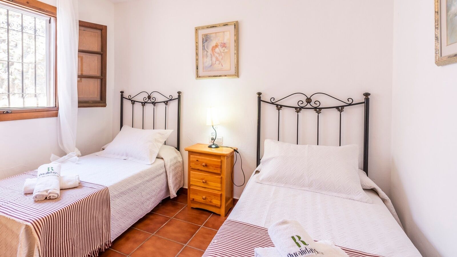 Una habitación con dos camas individuales, una mesita de noche de madera, una lámpara de pared y una ventana con cortinas.