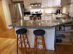 Una cocina con encimera de granito, electrodomésticos de acero inoxidable, taburetes de madera y una estantería con libros y objetos decorativos.
