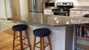 Una cocina con armarios blancos, electrodomésticos de acero inoxidable, encimera de granito y dos taburetes de madera.