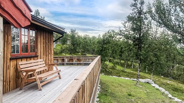 A wooden cabin with a deck and bench, surrounded by trees and a grassy area.