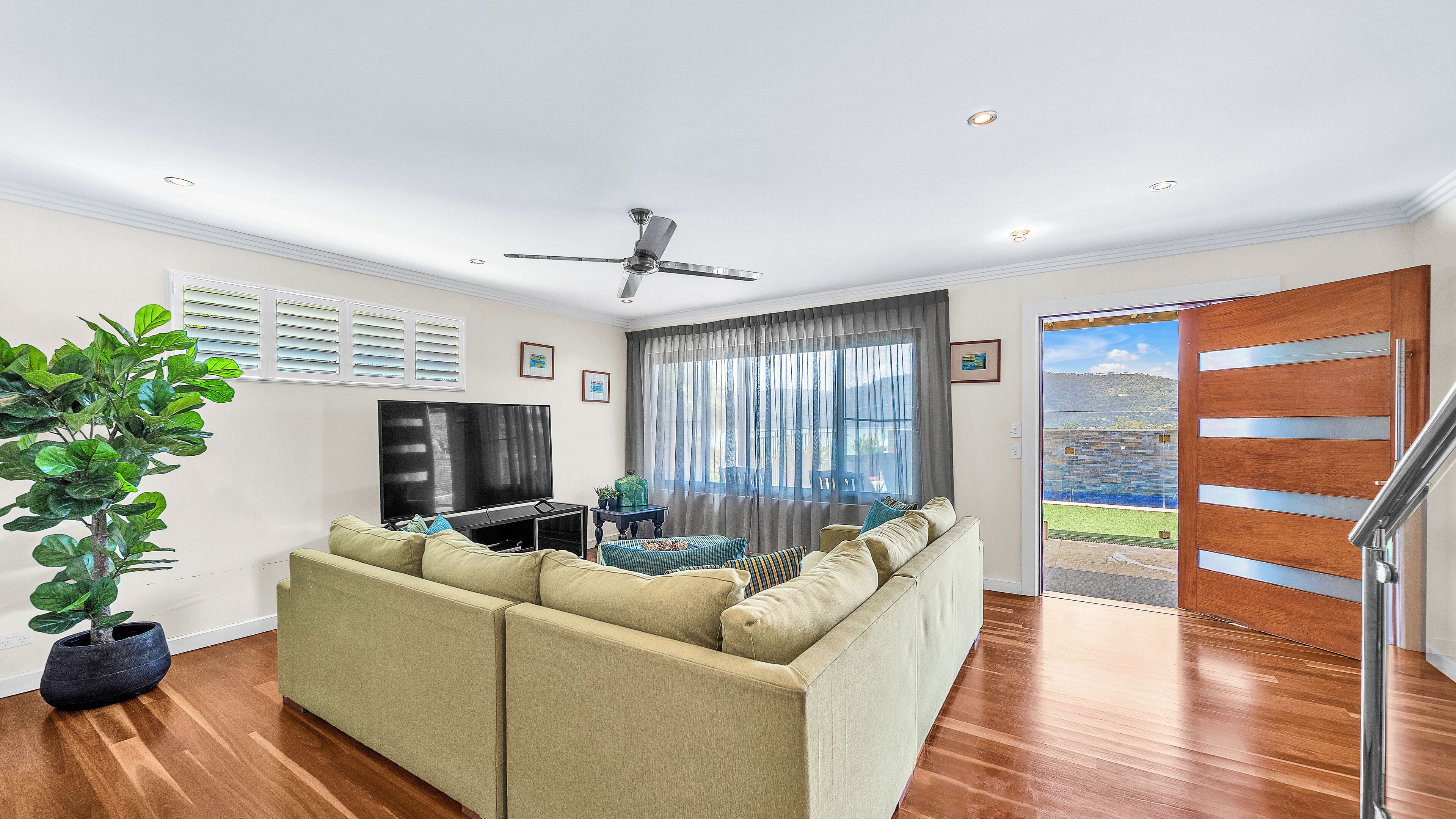 A modern living room with a sectional sofa, a flat-screen TV, a ceiling fan, and a large window with curtains.