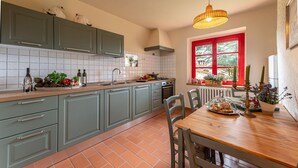 Une cuisine avec des meubles de couleur grise, un plan de travail en bois et une table à manger avec des chaises. Il y a une fenêtre avec des volets rouges et un luminaire suspendu.