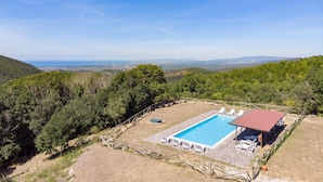 Une piscine entourée de pierres et de chaises longues blanches, surmontée d’un toit rouge, le tout se fondant dans un paysage de collines verdoyantes et offrant une vue lointaine sur la mer.