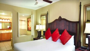 Ein Hotelzimmer mit einem großen Bett, einem hölzernen Kopfteil, einem Nachttisch, einem Spiegel und einem im Hintergrund sichtbaren Badezimmer.