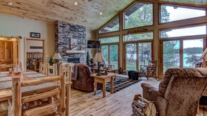 A spacious living room with a stone fireplace, wooden furniture, and large windows offering a view of the outdoors.