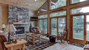 A cozy living room with a stone fireplace, wooden furniture, and large windows offering a view of the outdoors.
